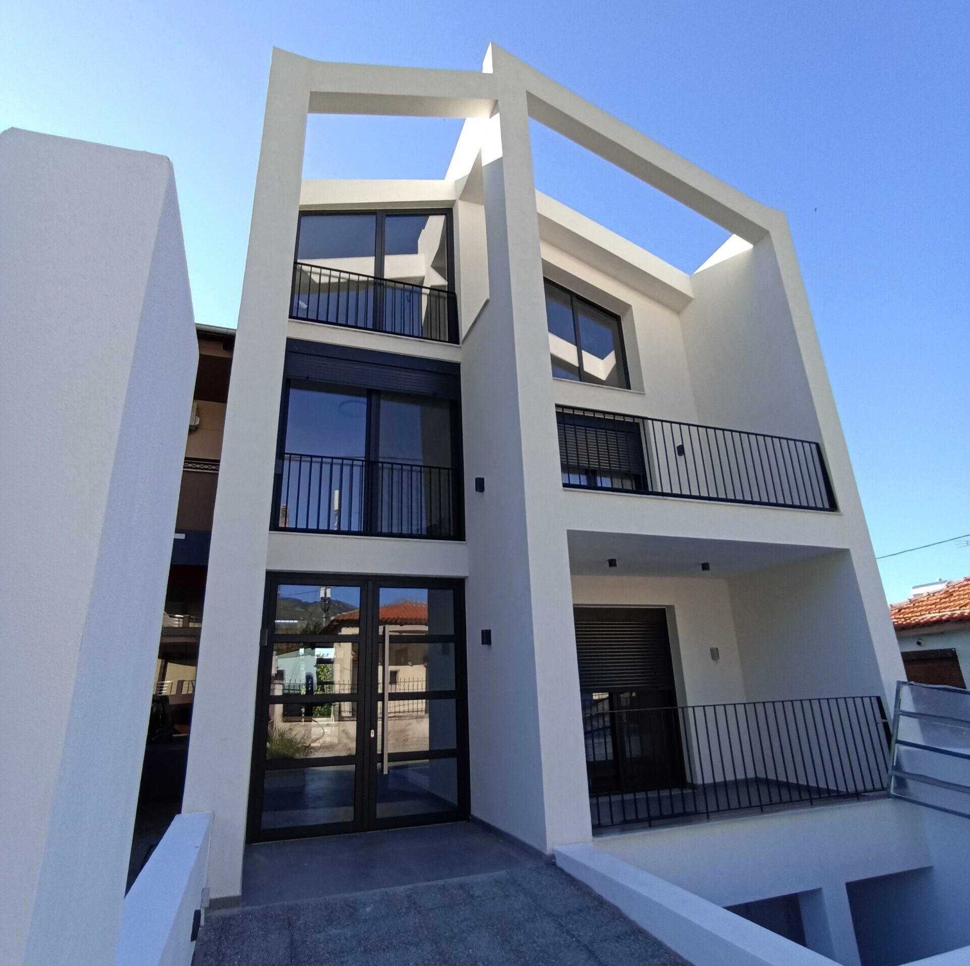 A modern white three-story house with large glass windows, black railings, and wooden pergola accents.