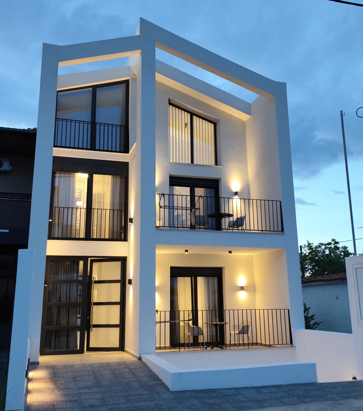 Alternative view of a modern white three-story house with large glass windows, black railings, and wooden pergola accents.